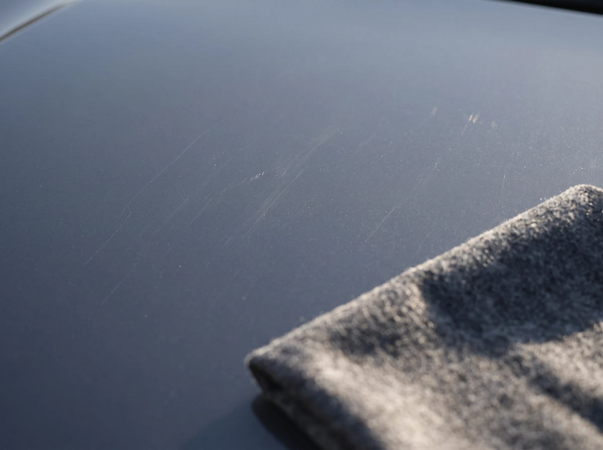 Close-up of fine light scratches on dark gray car paint, with a folded NanoPolix Nano Sparkle Cloth resting in the corner